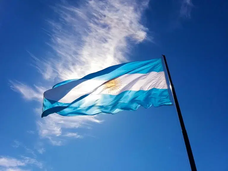 Le drapeau argentin flottant sous un ciel bleu clair avec des nuages légers, montrant ses bandes bleu ciel et blanche avec un soleil doré rayonnant au centre.