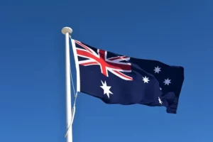 Le drapeau de l’Australie flottant sur un mât blanc sous un ciel bleu clair, mettant en avant l’Union Jack et les étoiles de la constellation du Sud.